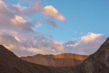 Dramatic landscape with sunlit wide sharp mountain ridge under clouds in sunset colors at changeable weather. Atmospheric mountain scenery with large sharp rocks on ridge top under sunset cloudy sky.