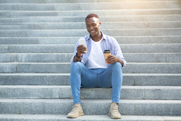 Urban lifestyle. Attractive black man with coffee to go using mobile phone on stone stairs outdoos