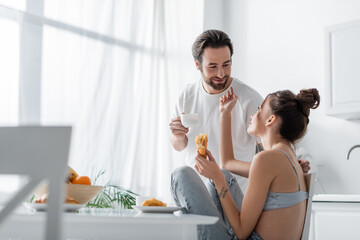 tattooed young woman holding fresh croissant and reaching happy boyfriend with cup.