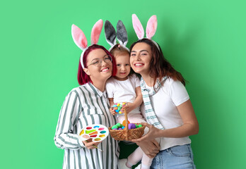 Young lesbian couple with little daughter, Easter eggs and paints on color background