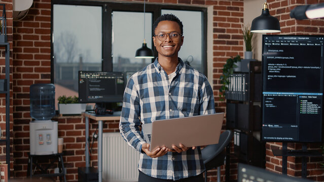 Portrait of african american programer standing working on laptop looking up and smiling at camera. Coder holding portable computer next to coworker computing big data in it agency office.