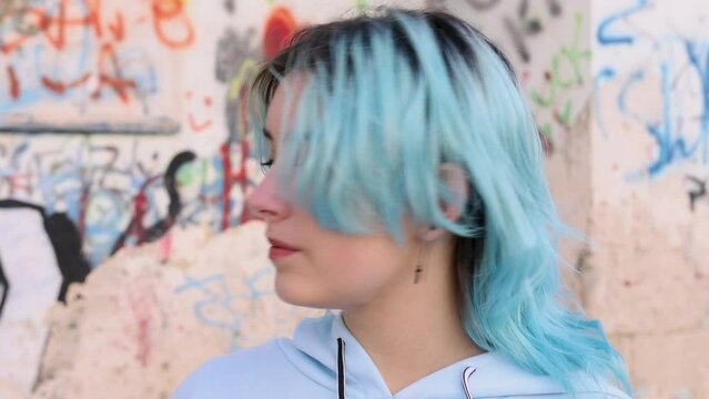 Serious Teenager in light blue oversize hoodie turn left. Blue haired teen girl staying outdoor against graffiti wall and looking into camera. Head shot