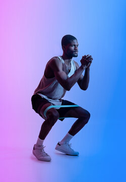 Full length of young black sportsman doing squats with fitness resistance band in neon lighting. Strength workout