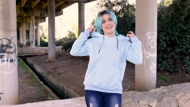 Smiling teenager wearing hoodie and staying against bridge pillars. Blue haired teen girl stays outdoors against bridge pillars. Clothing mockup