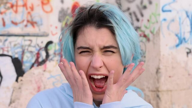 Serious Teenager in light blue oversize hoodie screaming with hands near head. Blue haired teen girl staying outdoor against graffiti wall and looking into camera. Head shot