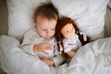 Fototapeta na wymiar A cute Caucasian baby with a doll and with a thermometer, a sick child lies in bed with a toy, bed rest during illness. Comfort and play in bed
