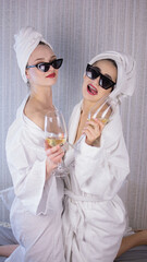 Front view of two smiling female friends with towels and in bathrobes sitting on bed and doing pedicure. Cute happy young girls doing beauty procedure and drinking wine, enjoying time together.