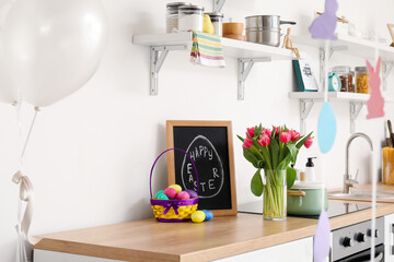 Chalkboard with text HAPPY EASTER, eggs and tulips on table in kitchen