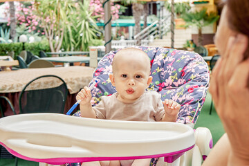 Small boy sits in baby armchair of outdoor restaurant under mother care. Child plays with blue pen against colorful and blooming garden plants