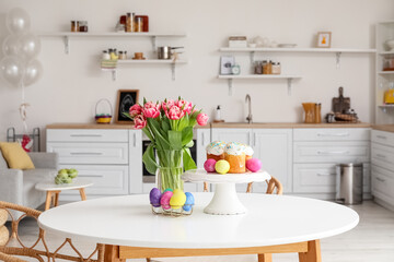 Stand with Easter cakes, eggs and tulips on dining table in kitchen