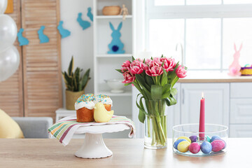 Stand with Easter cakes, eggs, burning candle and tulips on counter in kitchen