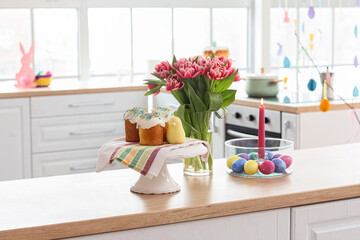 Stand with Easter cakes, eggs, burning candle and tulips on counter in kitchen