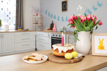 Fototapeta na wymiar Easter cake, candle, eggs, cookies, picture and vase with tulips on counter in kitchen