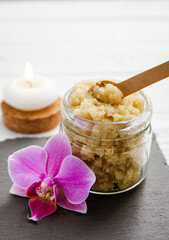 Yellow color handmade sugar body scrub in glass jar. Burning candle and pink orchid blossom for decoration on white studio background. Copy space.