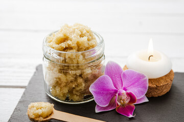 Yellow color handmade sugar body scrub in glass jar. Burning candle and pink orchid blossom for decoration on white studio background. Copy space.