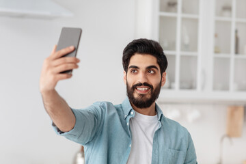 Young arab man holding smartphone, taking selfie or making video call, standing in kitchen interior.