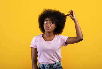 Upset african american lady touching and showing her curly hair on yellow background, studio shot