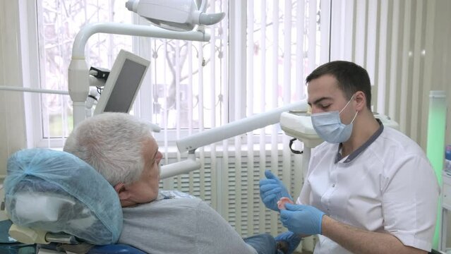 Male dentist advises elderly patient about removable dentures in dental clinic. Doctor communicates with patient. Dentist explaining artificial teeth to senior man. Healthcare and dentistry business