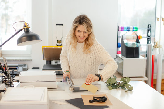Professional smiling blond woman in sweater worker using printer cutter in workshop. Print office. Carving sheets paper 