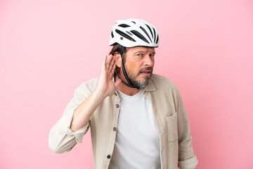 Senior dutch man with bike helmet isolated on pink background listening to something by putting hand on the ear