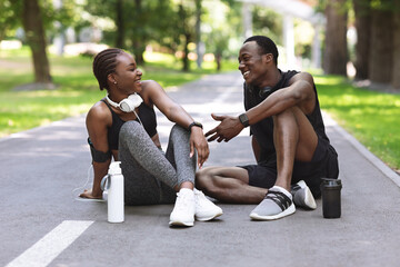Joyful Black Couple Relaxing After Training Outdoors, Sitting On Path In Park