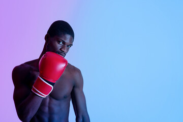 Determined young black boxer with bare torso wearing fighting gloves, looking at camera in neon...