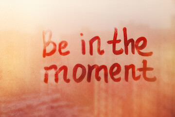 Hand drawn message Be in the moment on foggy glass on suset window