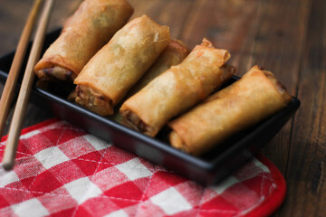Fried spring rolls served in a black square plate with sliced tomatoes on a brown wooden table.