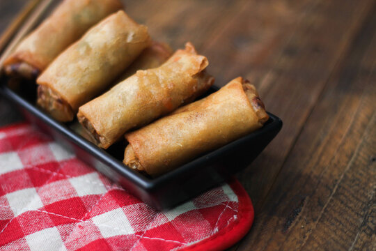 Fried spring rolls served in a black square plate with sliced tomatoes on a brown wooden table.