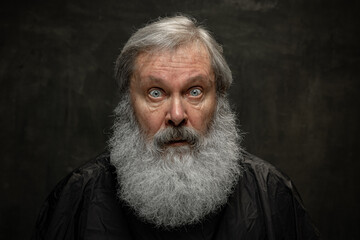 Obraz premium Dramatic portrait of emotional senior bearded man getting beard grooming isolated on dark vintage background. Concept of emotions, fashion, beauty, self-reinvention