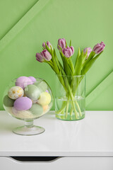 Different Easter eggs and vase with tulips on chest of drawers near color wall