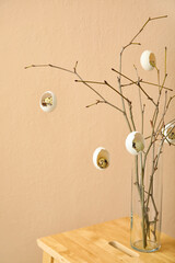 Tree branches decorated with cracked Easter eggs in vase on wooden stool near color wall