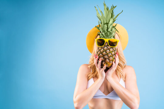 Portrait of comic funny girl with slender figure in white two pieces swimsuit isolated on light blue background. Young blond woman is holding pineapple in glasses. Pop art concept. Summer mood.