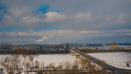 An urban landscape with a frozen and snow-covered river on the background of a big city. A road bridge, a modern stadium and an ancient cathedral. The spring blue sky emerging through the clouds.