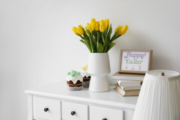 Tulips in vase, frame with text HAPPY EASTER, books and cakes on table near light wall