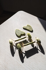 Two green ceramic jade roller and gouache scraper on a white wooden background, table, sunlight . Facial massage, beauty, youth, anti-wrinkle, beauty, home morning daily care. Anti-aging.
