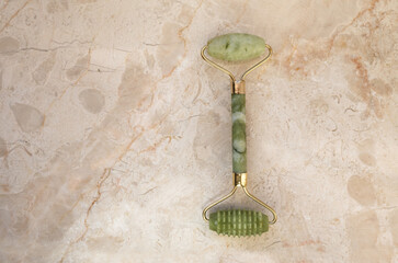 Green ceramic jade roller and gouache scraper on a beige marble  background, table. Facial massage,...