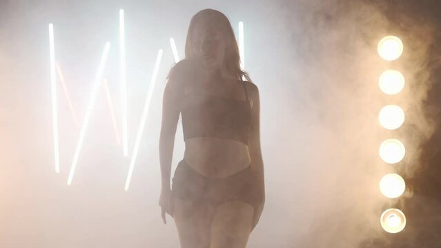 Artistic young woman dancing in backlit fog smoke indoors. Confident graceful Caucasian female dancer rehearsing modern performance with ballet movements. Handheld effect choreography. Neon light