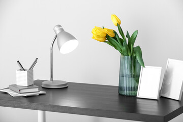 Vase with tulips, glowing lamp and stationery supplies on table near light wall