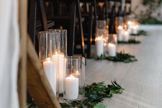 Lit candles in transparent glass jars on the floor near the row of chairs A glass vases with burning candles inside and greenery decoration Cropped photo Close up 