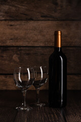 Bottle of red wine with two empty glasses on rustic wooden background, suitable for wine tasting events.