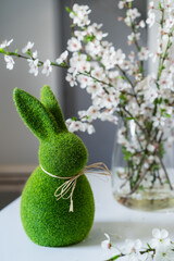 Green easter bunny with blooming flowers on tree branch in the vase on the white table. Happy Easter spring vertical card. Selective focus