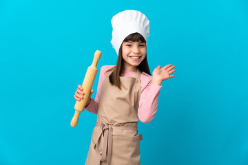 Little girl holding a rolling pin isolated on blue background saluting with hand with happy expression