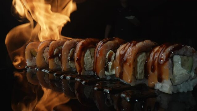 Philadelphia sushi roll with salmon and unagi sauce roasted on fire. Fire envelops set of sushi on black background close up. Sushi roll grill with salmon, rice, avocado and cheese. Slow motion.