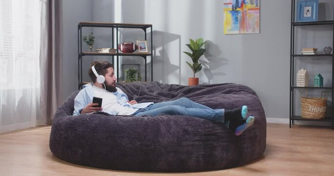Full size portrait of adult man laying back on sofa in casual clothes. Man listening to rythmic music. Man chilling on couch and enjoying. Happiness concept. Slow motion video.