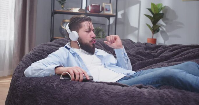 Close up portrait of adult man laying back on sofa in casual clothes. Man listening to rythmic music and making dancing moves. Man chilling on couch and enjoying. Happiness concept. Slow motion video.