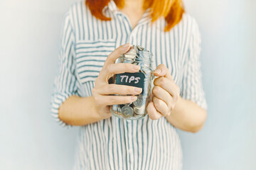 Glass bottle with silver coins in women's hands. Sticker with inscription on money jar. Redhead girl n white and black striped shirt holds her tips pot.