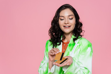 joyful woman in green tie dye blazer looking at credit cards and smiling isolated on pink.