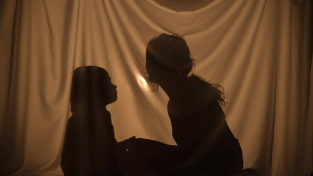Unrecognizable woman and boy sharing secrets and hugging. Mom and little son spend leisure together, sitting behind curtain at home. Puppet theater of shadows, play of shadow and light. Close up.