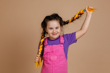 Beautiful, little girl, pulling kanekalon pigtails of yellow color, looking at camera with gentle smile, wearing pink jumpsuit and purple t-shirt on beige background.